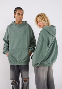 Felpe verde oversize con tasche frontali e logo ricamato "UR TRN", abbinate a jeans neri strappati. Tessuto morbido con design casual.