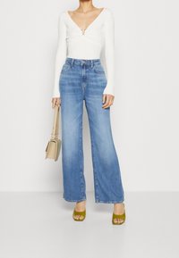 Biała bluzka z długim rękawem i głębokim dekoltem w kształcie litery V, ozdobiona zapięciem, w połączeniu z jasnoniebieskimi spodniami o szerokich nogawkach. Modelka trzyma beżową torebkę i ma na sobie zielone szpilki.