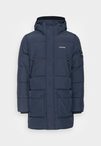 Granatowa pikowana kurtka z kapturem, wyposażona w kieszenie na piersi na zamek błyskawiczny, zapięcia na zatrzaski oraz akcenty z logo na piersi i rękawie.