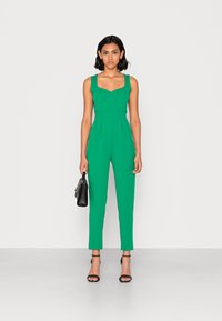 Macacão verde sem mangas com corpete ajustado, pernas afuniladas e bolsos laterais. Modelo segura uma mala preta, usando sandálias pretas com tiras.