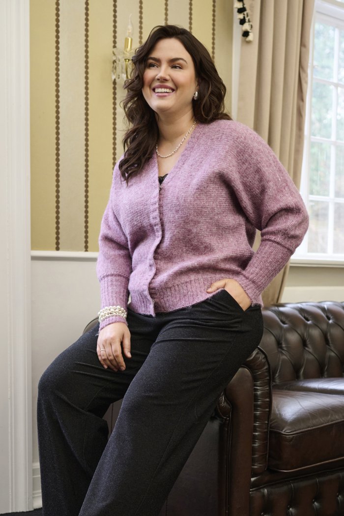 Cardigan en tricot rose doux avec un tissu texturé, présentant un col en V, une fermeture à boutons et des poignets côtelés, associé à un pantalon sombre.