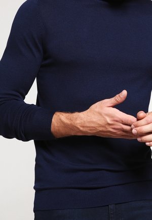 Mężczyzna w granatowej koszuli z długim rękawem, z rękami splecionymi, z widocznymi żyłami i jasnym odcieniem skóry.