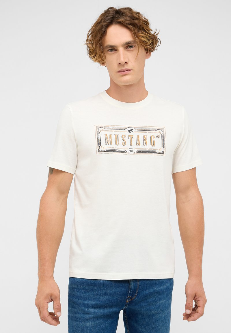Young man with curly hair wearing white Mustang logo t-shirt and blue jeans, standing against plain light background.