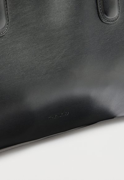 Κοντινή λήψη από λείο μαύρο δερμάτινο τσάντα tote με ραμμένες λαβές και διακριτικό εξώγλυφο εμπορικό σήμα "The Row" κοντά στον πάτο.