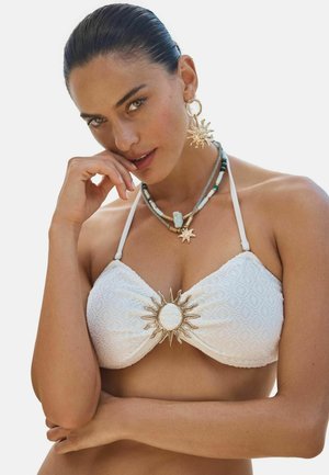 Nő hátrafésült hajjal fehér, texturált bikini felsőt visel arany nap kitűzővel, nap formájú fülbevalókkal és rétegzett gyöngyös nyakláncokkal.