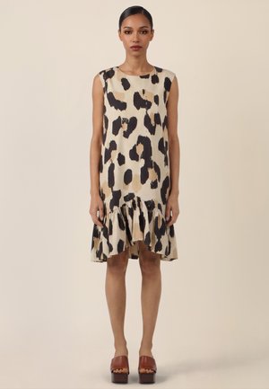 Frau steht und trägt ein ärmelloses beiges Kleid mit schwarzen und braunen abstrakten Flecken und einem Volant-Saum, kombiniert mit braunen Plateau-Sandalen.