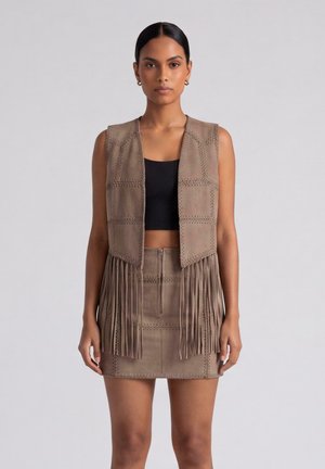 Donna che indossa un gilet in camoscio marrone con frange e mini gonna abbinata, abbinata a un crop top nero, in piedi davanti a uno sfondo semplice.