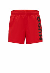 Red swim shorts with an elastic waistband, featuring a large black "HUGO" logo printed vertically on the left side. Made from lightweight material.
