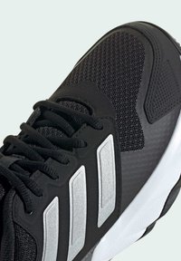 Zapatilla deportiva negra con materiales de malla y sintéticos, que presenta tres franjas plateadas, paneles texturizados y una suela de caucho blanca.