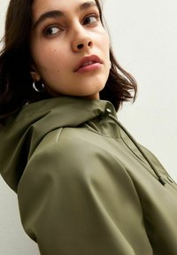 Chaqueta impermeable verde oliva con capucha, que presenta una textura suave, cordón elástico y un botón cerca del cuello para un cierre seguro.