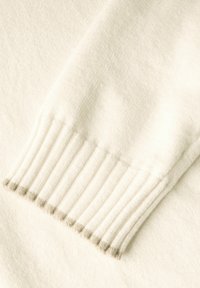 Maniche di un maglione color crema realizzate in tessuto morbido, con texture a costine vicino al polsino e sottili accenti grigi sui bordi.