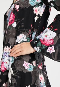 Mustakukkainen mekko, jossa on kirkkaan vaaleanpunaisia, valkoisia ja sinisiä kukkia, pitkät hihat ja sileä, kevytrakenne.