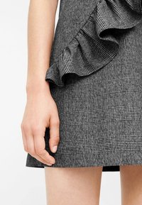 Grauer karierten Rock mit einer gerafften Akzentuierung an der Seite. Strukturierter Stoff; eng anliegendes Design; Hand ruht auf dem Saum.