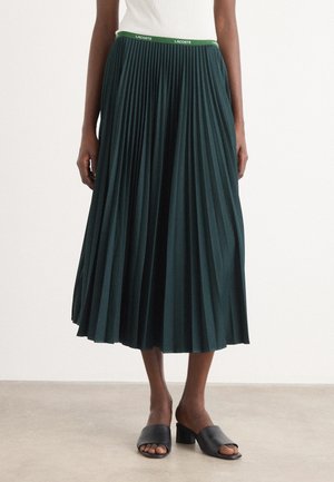 Jupe midi plissée verte en tissu texturé, avec une ceinture de marque contrastante, associée à des chaussures à talons ouvertes noires.