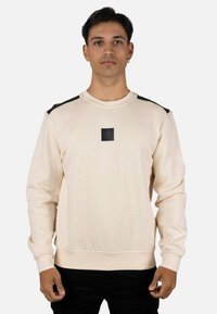 Sudadera de color beige claro con detalles negros en los hombros y un parche de logo central. Presenta cuello redondo y puños acanalados. Textura de tela suave.