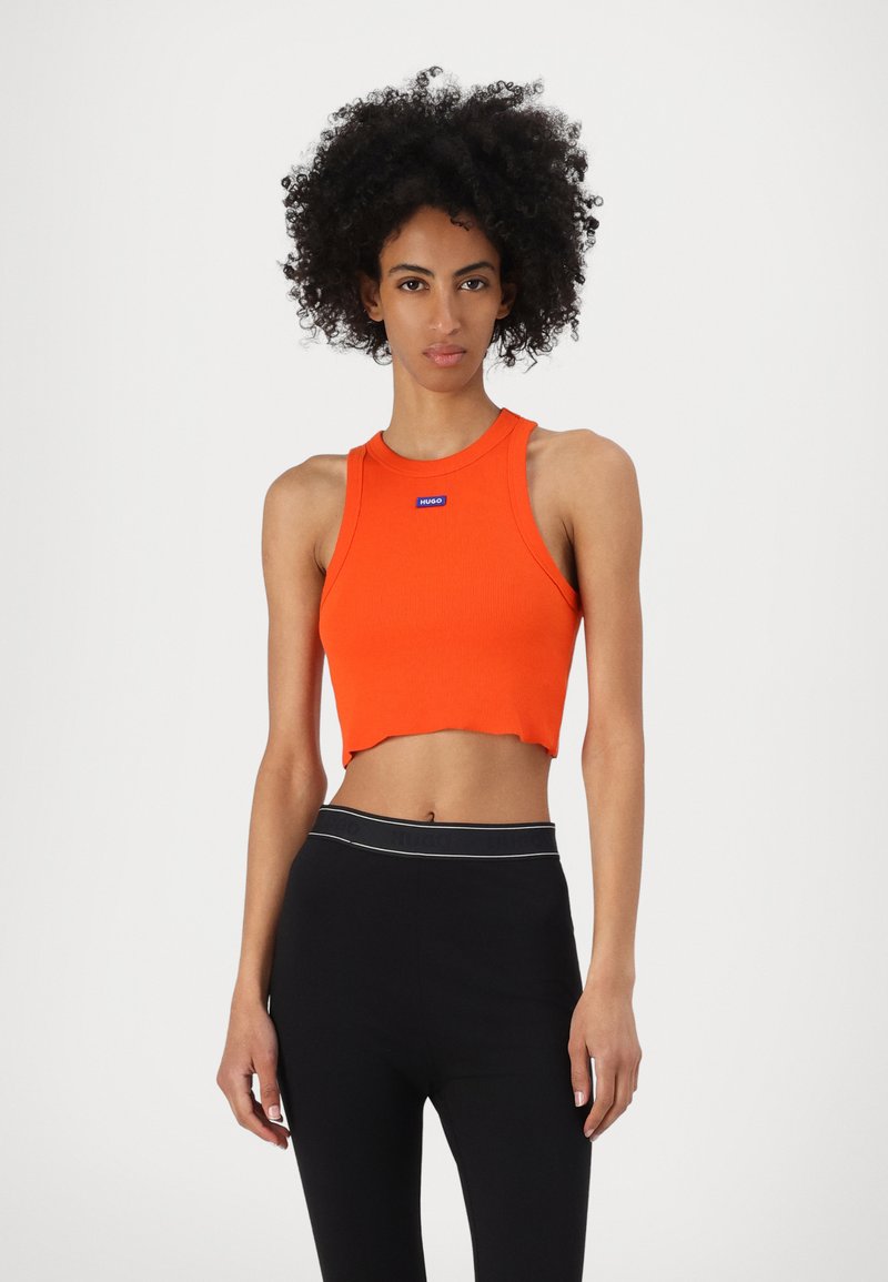 Crop top orange à côtes avec un col rond et un ourlet courbé. Présente une étiquette logo rectangulaire bleue à l'avant. Porte avec des leggings noirs.
