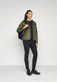 Casaco acolchoado verde-oliva, top preto, calças cargo pretas com bolso e ténis pretos. O casaco tem um emblema Fjäder.