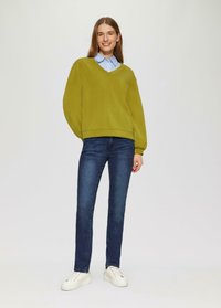 Lächelnde Frau steht in einem grünen V-Ausschnitt-Pullover über einem blauen gestreiften Hemd, dunkle Jeans und weiße Sneakers vor einem schlichten Hintergrund.