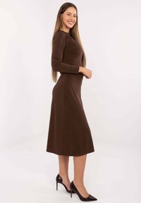 Brązowa, dopasowana sukienka z długim rękawem o długości do połowy łydki, charakteryzująca się gładką fakturą i subtelnymi akcentami na ramionach, zestawiona z czarnymi szpilkami.