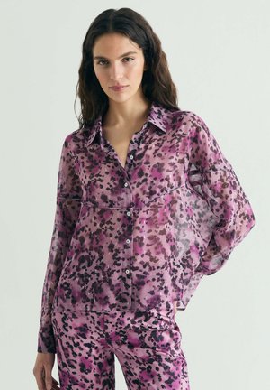 Mujer con blusa transparente de botones, morada con manchas negras, y pantalones a juego, posando con una mano en la cadera frente a un fondo liso.