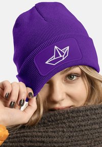 Purple knit beanie with a textured pattern and a white paper boat design on a rectangular patch. Wrapping a knitted brown scarf.