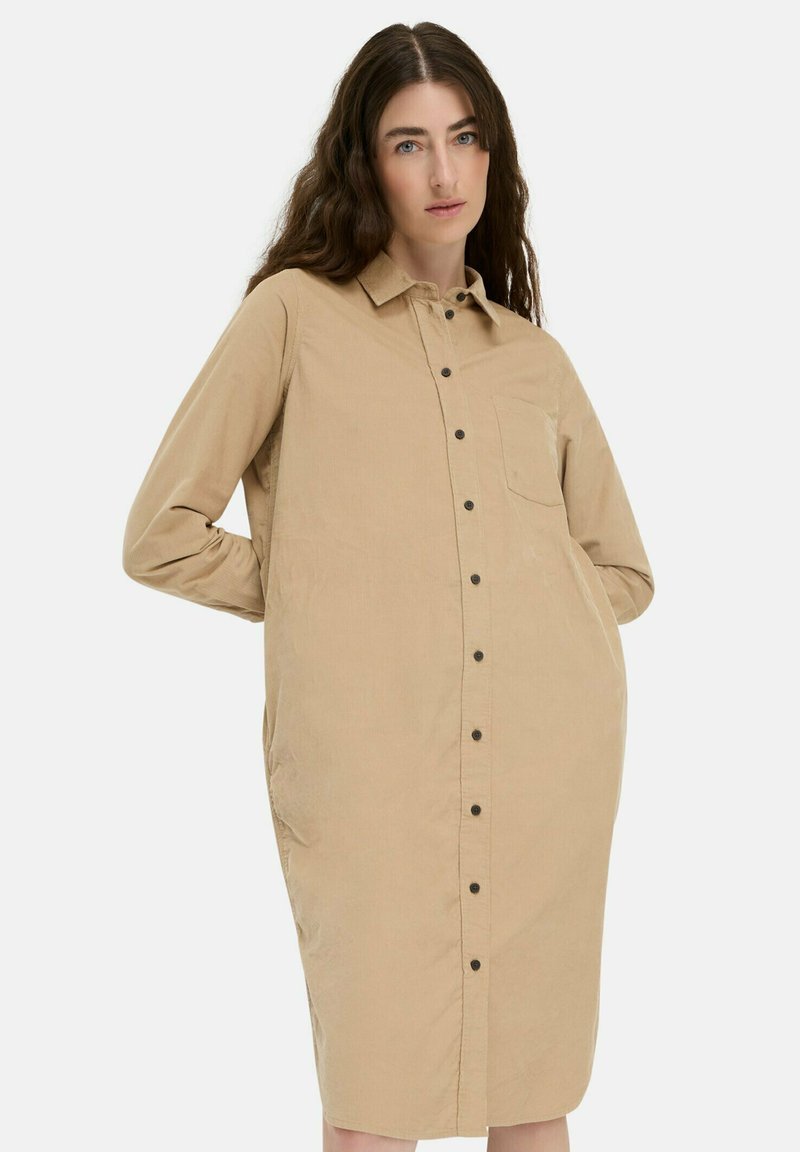 Frau mit welligem braunem Haar, die ein beiges, knöpfbares Kordhemdkleid mit langen Ärmeln und einer Brusttasche trägt, steht mit den Händen hinter dem Rücken.