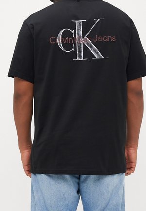 Schwarzes T-Shirt mit großen distressed "CK" Buchstaben und "Calvin Klein Jeans" Schriftzug am oberen Rücken, getragen mit hellblauen Jeans.