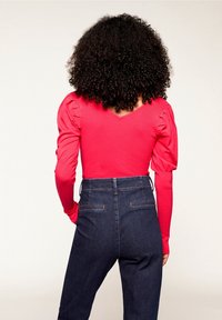 Persona con capelli ricci che indossa un top lungo rosa brillante con spalle gonfie e jeans blu scuro a vita alta, di spalle.