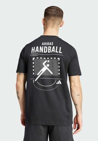 Camiseta negra de Adidas con un gran gráfico blanco que ilustra balonmano, con la palabra "HANDBALL" y diseños geométricos en la parte posterior.