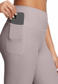 Primo piano di una mano che ripone uno smartphone nella tasca laterale di leggings mauve a vita alta con righe, indossati all'altezza dell'anca.