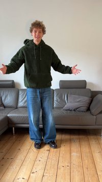 Grön fleecetröja med huva och framficka, lösa blå jeans och mörka skor. Står med armarna utsträckta i ett vardagsrum.