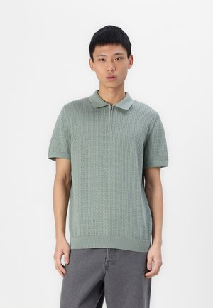 Hellgrünes Strick-Polohemd mit Reißverschluss, kurzen Ärmeln und strukturiertem Muster. Gerippter Kragen und Saum, kombiniert mit grauen Jeans.