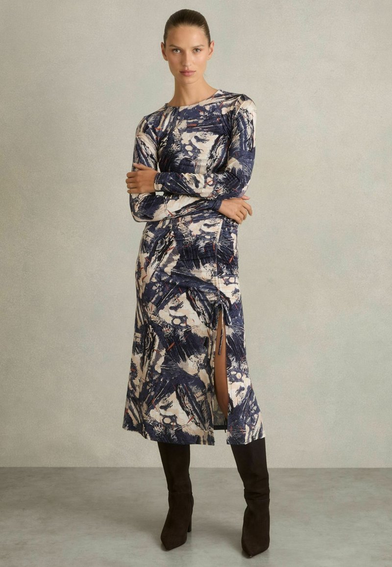 Vestido de manga larga con estampado abstracto en azul, blanco y rojo; de longitud media con abertura lateral. La tela tiene una textura suave. Se lleva con botas altas de rodilla oscuras.