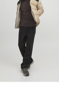 Giacca puffer beige con accenti neri, maglione marrone e pantaloni neri plissettati. Sneakers grigie completano l'outfit. Design semplice e funzionale.