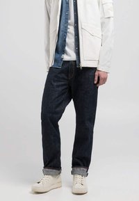Jeans de mezclilla oscura con un corte relajado, combinados con una chaqueta blanca clara, superpuesta sobre una camisa azul, y zapatillas blancas.