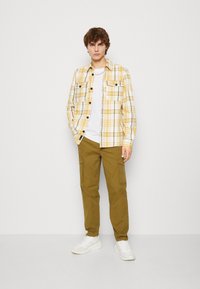 Chemise à carreaux jaune et blanche avec deux poches poitrine, portée par-dessus un t-shirt blanc. Un pantalon cargo kaki et des baskets blanches complètent la tenue.