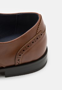 Zapato de cuero marrón con un acabado liso, que presenta perforaciones decorativas, una suela de goma negra contrastante y acentos cosidos a lo largo del borde.