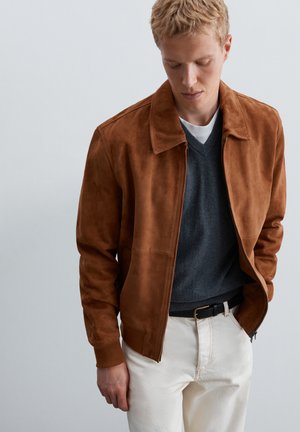 Jeune homme portant une veste en daim marron, un pull col en V gris foncé sur une chemise blanche, un pantalon blanc et une ceinture noire, regardant vers le bas, les mains dans les poches.