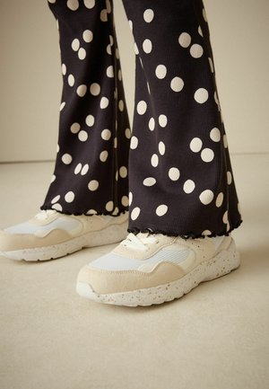 Beige och vita sneakers med strukturerade ovandelar och fläckiga sulor, matchade med svarta byxor med stora vita prickar.