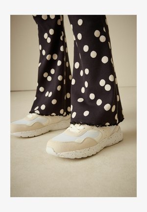 Smėlio spalvos ir baltos sportbačiai su tekstūruotu viršumi ir dėmėtomis padėlėmis, derinami su juodomis kelnėmis su didelėmis baltomis taškelėmis.