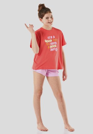 Coral t-shirt with colorful text, paired with light pink shorts. Casual fit with short sleeves and a round neckline. Barefoot pose.