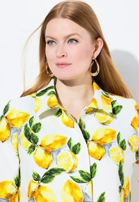 Camicia bianca con una vivace stampa di limoni e foglie; colletto strutturato, dettagli con bottoni e superficie in tessuto liscia. Orecchini a cerchio dorati in vista.