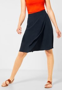 Námořnická zavinovací sukně s elastickým pasem, vyrobená z lehké tkaniny, v kombinaci s hnědými plochými sandály. Hladká textura a casualní design.