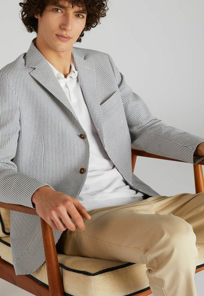 Pasiasty blezer w jasnym niebieskim i białym kolorze z bawełny, z dwiema brązowymi guzikami, w połączeniu z białą koszulą i beżowymi spodniami. Siedzi na drewnianym krześle.