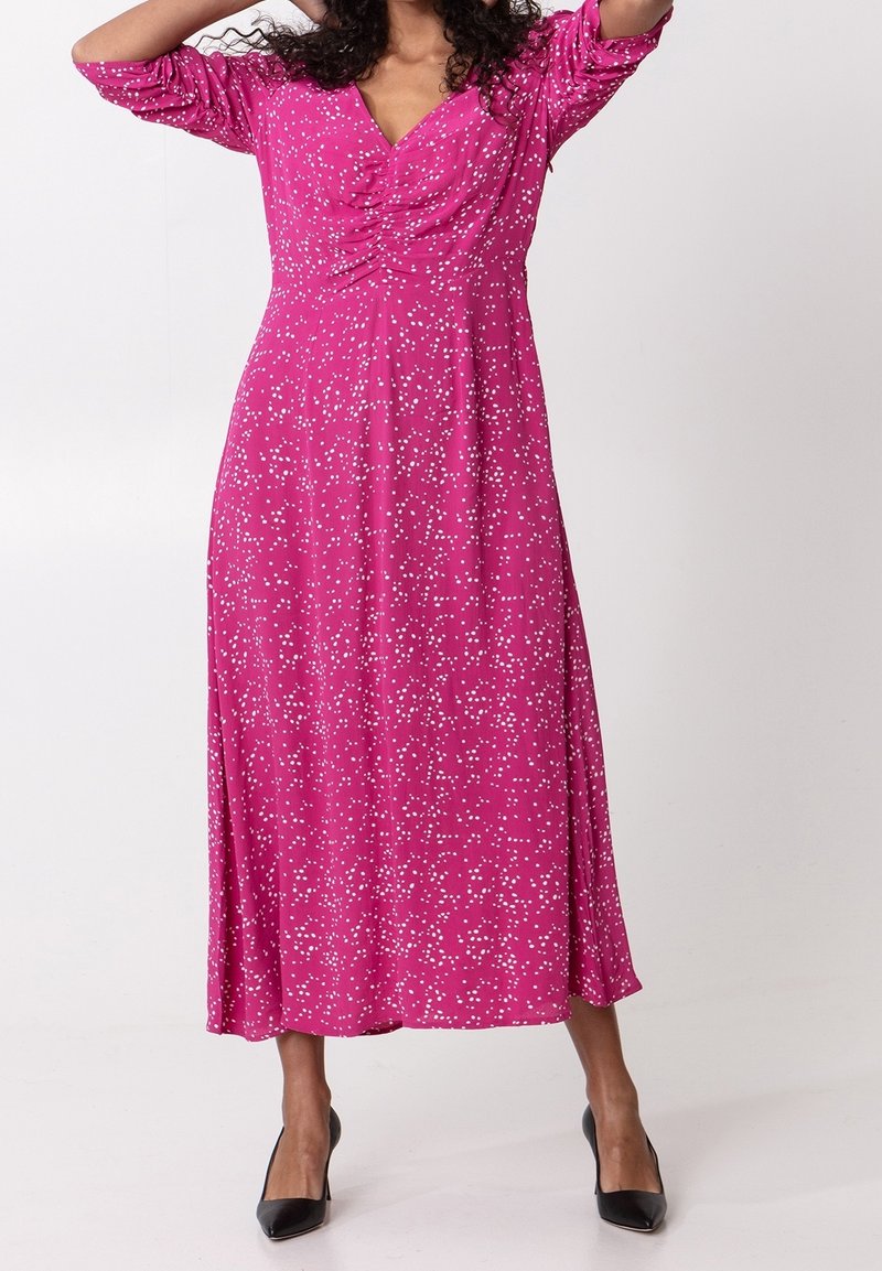 Femme portant une longue robe rose à pois blancs, avec un col froncé, des manches trois-quarts, et des talons noirs pointus, debout sur un fond blanc.