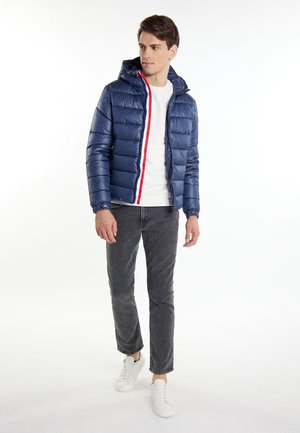 Navy gepolsterte Jacke mit Kapuze, ausgestattet mit einem roten und weißen Reißverschluss, kombiniert mit grauen Hosen und weißen Turnschuhen für einen lässigen Look.