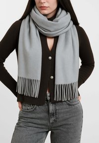 Sciarpa in lana grigia chiara con estremità frangiate, indossata sopra un cardigan marrone scuro con bottoni argentati, esposta su uno sfondo uniforme.