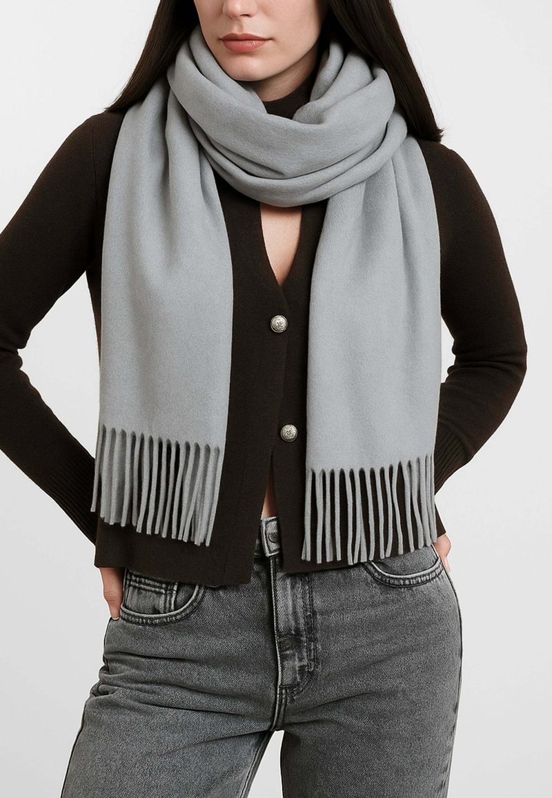 Sciarpa in lana grigia chiara con estremità frangiate, indossata sopra un cardigan marrone scuro con bottoni argentati, esposta su uno sfondo uniforme.