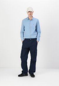 Light blue button-up shirt, navy cargo pants with side pockets, and a beige cap. Black shoes complete the outfit. Minimalist design.