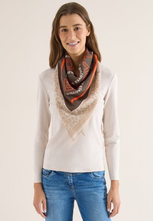 Femme portant une chemise à manches longues beige clair, un jean bleu, et un grand foulard triangulaire aux motifs orange, marron et beige autour du cou.